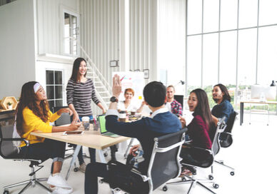 Group of multiracial young creative team talking, laughing and brainstorming in meeting at modern office concept. Female standing and man raising hand for sharing while sitting together in rear view.